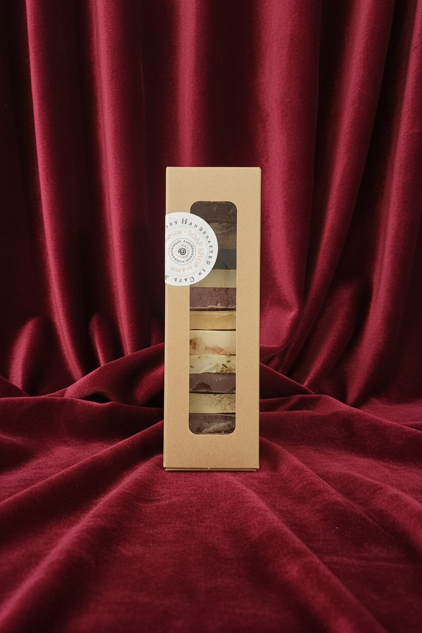 A rectangular kraft paper box containing a dozen guest sized bars of handmade soap, with a round white sticker on the left side that reads 'Handcrafted in Cape May'. The box is placed on a rich velvet fabric background.