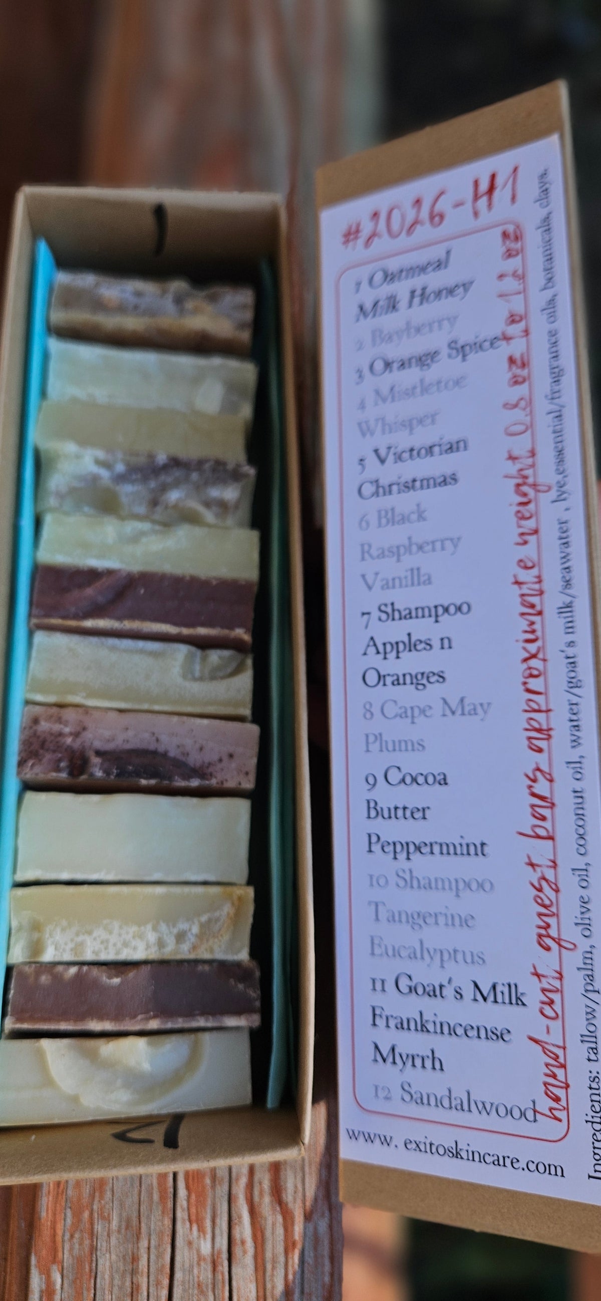 A cardboard box of handmade soap bars, with the lid open to show a handwritten list of ingredients on the inside. The soaps are arranged in vertical columns, showing various colors like blue, green, brown, beige, and white.