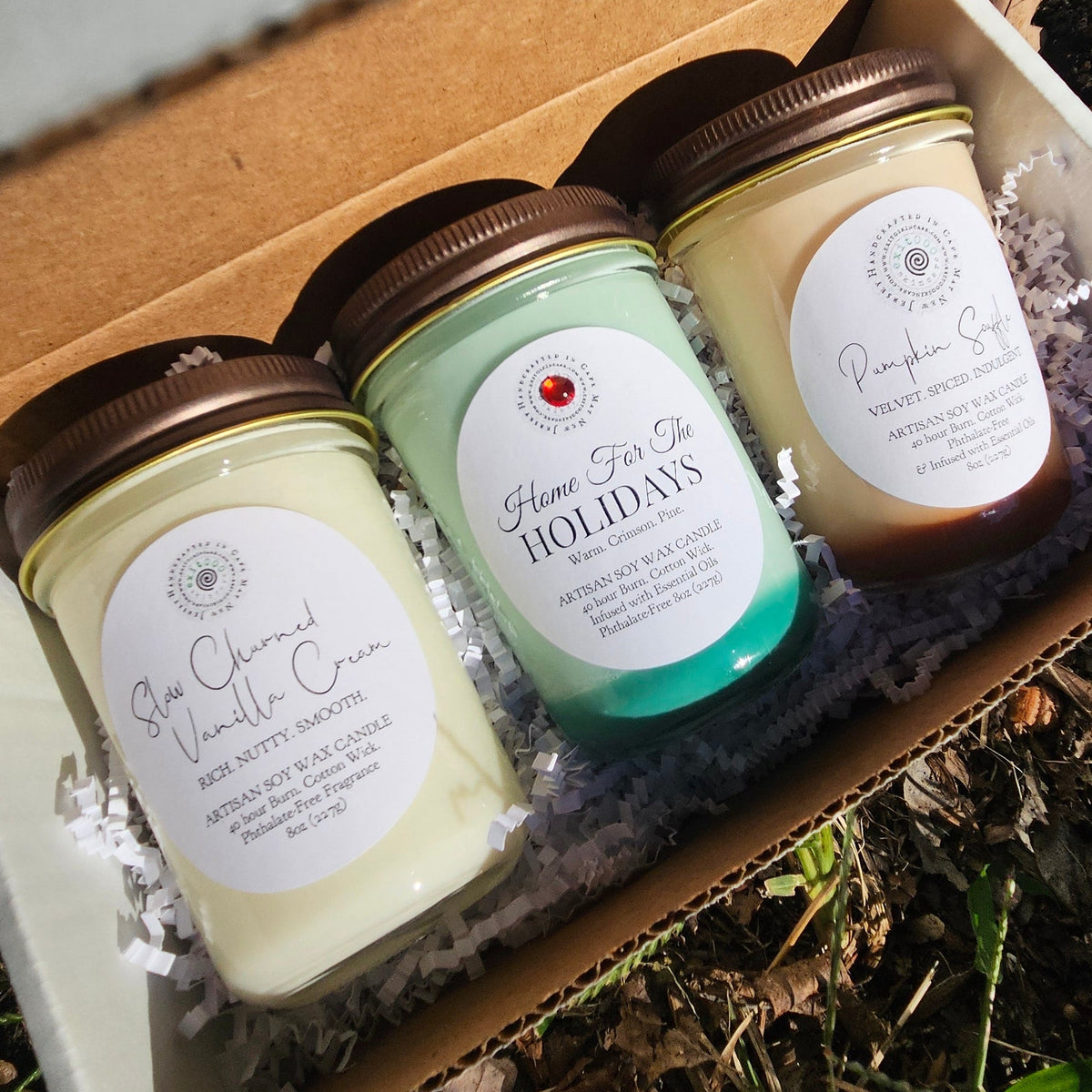 Three small jars of body butter or soap with white labels are arranged inside an open cardboard box on a bed of natural wood chips and dried leaves outdoors.